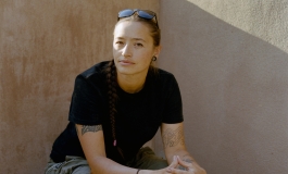 Photo of a person sitting next to a pueblo wall in casual clothes, peering at the camera