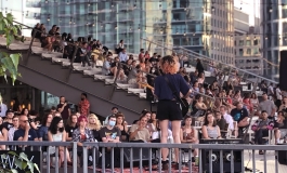 An audience sitting outside the ICA and on the grandstand watch a musical performer on a stage with their back to the camera