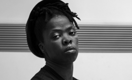 A portrait of artist Zanele Muholi with their head raised and arms crossed looking into the camera.