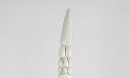 A tall, slender sculpture in white wood of an abstracted figure with carved rounded shapes at the top and a navel-like depression near the midpoint.