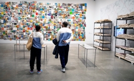 Two people in matching outfits look at wall of book installation while surrounded by glass desk cases
