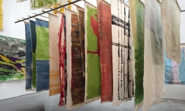 Unframed canvases painted with abstract shapes hang from a metal rack suspended from the ceiling in a gallery space.