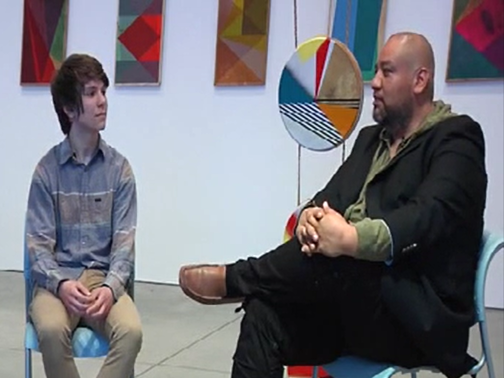 A light-skinned teenager with brown hair wearing a multicolored shirt and khaki pants sits across from a medium-skinned bald man wearing a green shirt, black pants and jacket, and brown shoes. They sit in front of art hanging on the walls and in the middle of the room with many colors and geometric and other patterns