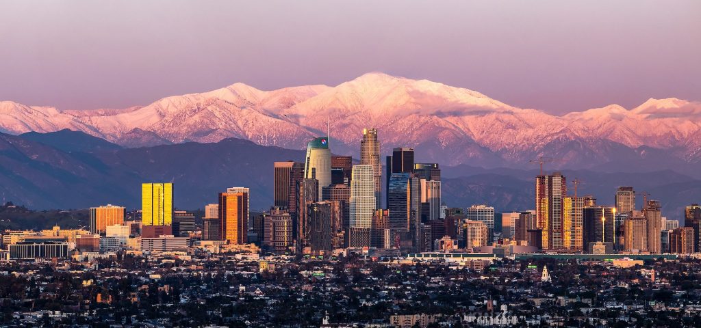 LA city skyline with tall buildings is set against snow-capped mountains at sunrise or sunset, with warm light reflecting off the skyscrapers and a gradient sky overhead.