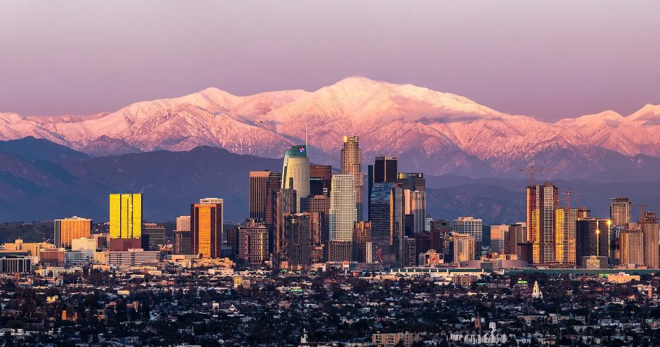 LA city skyline with tall buildings is set against snow-capped mountains at sunrise or sunset, with warm light reflecting off the skyscrapers and a gradient sky overhead.