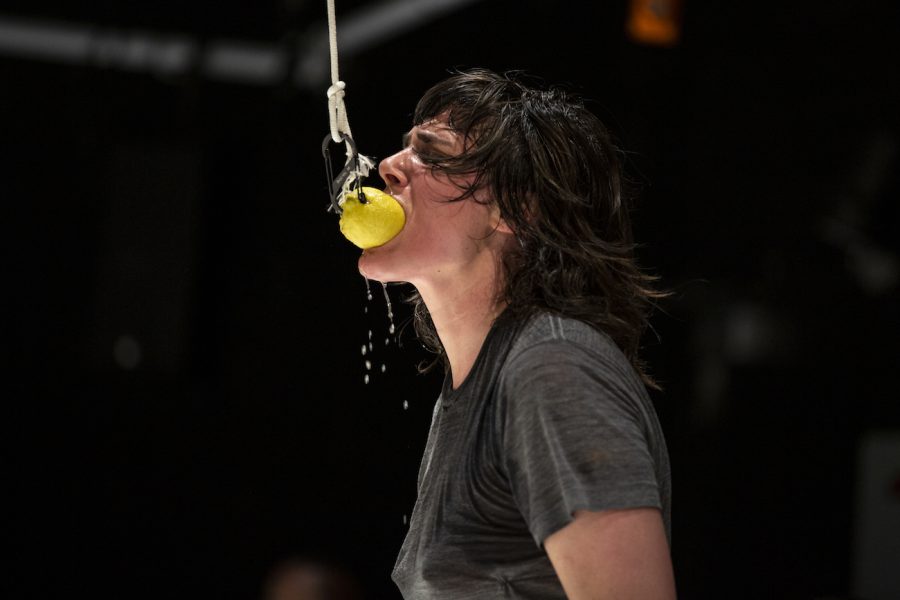 A dark-haired woman bites into a lemon hanging from a hook in a dark space. Juice drips off her chin.