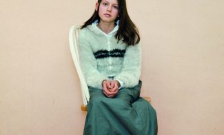 A color photograph of a light-skinned girl with long brown hair wearing a pale blouse and long green skirt sitting sideways on a chair to face the viewer.