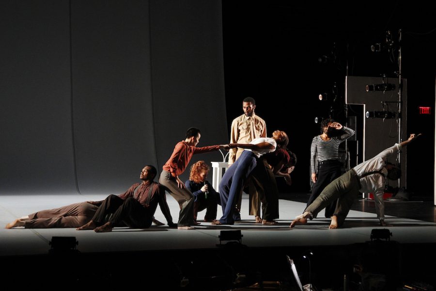 Nine dancers in plain, old-fashioned looking streetwear are posed across a dimly lit stage, some lying, some sitting, some standing.