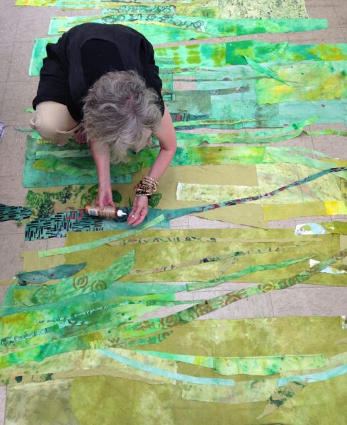 Fiber artist Merill Comeau kneeled down in her studio; she is putting glue in her hands and putting together strands of green cut-fabric to create a large scale collage.