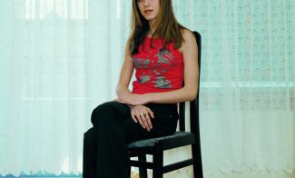 A color photograph of an light-skinned adolescent girl with long brown hair wearing a red tank and black pants sitting on a chair, positioned at an angle and gazing at the viewer.