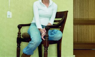 A color photograph of a light-skinned young woman wearing a white blouse and blue jeans, sitting sideways on a chair to face the viewer.