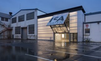 A building with a translucent front wall and garage-door like opening reading "ICA Watershed" seems to glow from within in an industrial area at dusk.