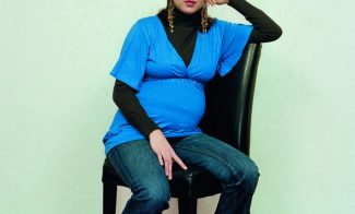 A color photograph of a pregnant light-skinned woman wearing a bright blue blouse and sitting with one arm propped on the back of a black chair, positioned at an angle and gazing at the viewer.