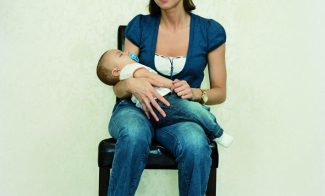 A color photograph of a light-skinned woman with short brown hair in a dark top, jeans, and white socks sitting in a chair with a baby in her arms, facing and gazing at the viewer.
