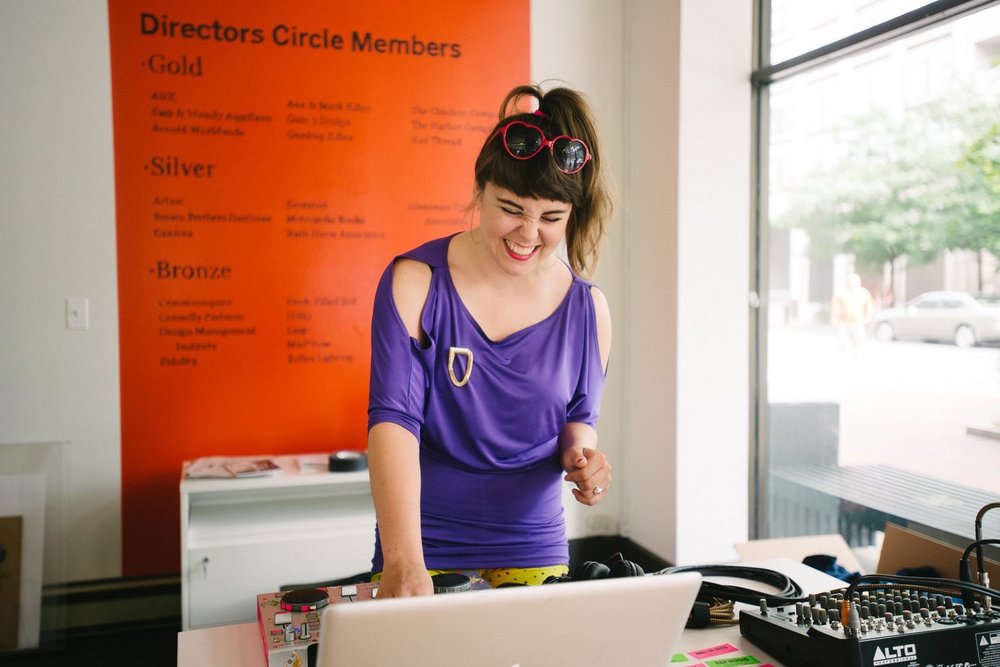 A DJ smiling over her laptop and soundboard equipment in a sunny, well-lit venue.