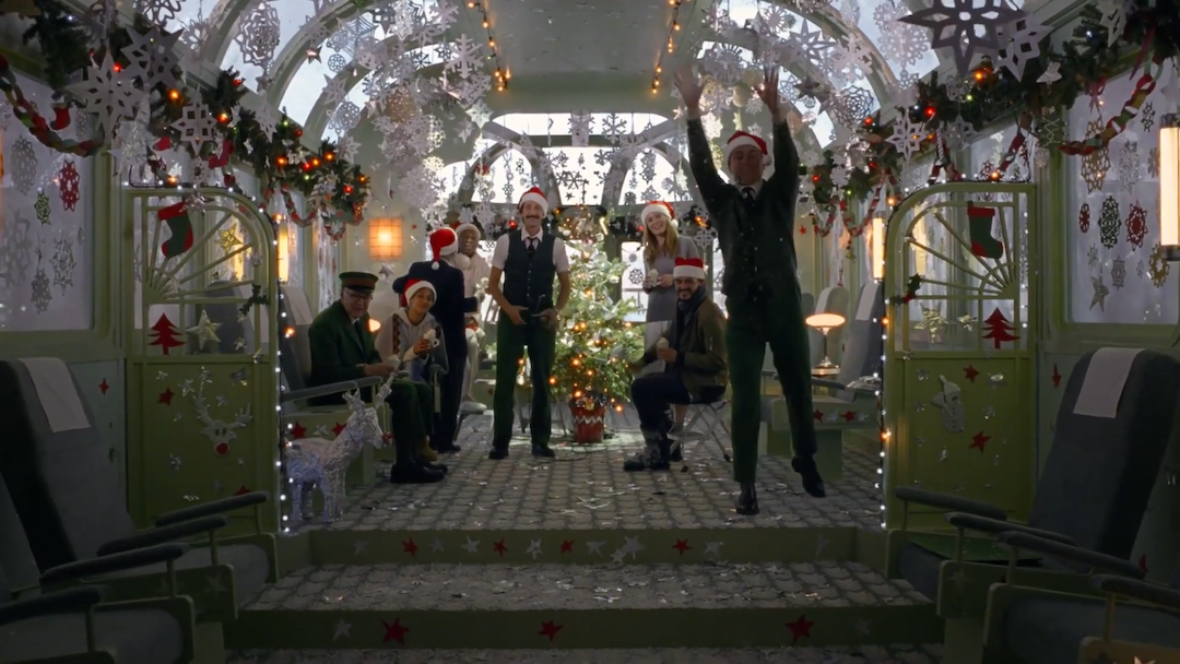 A group of people wearing Christmas hats, pedestrians and train conductors, looking to camera while in a festively decorated train set.
