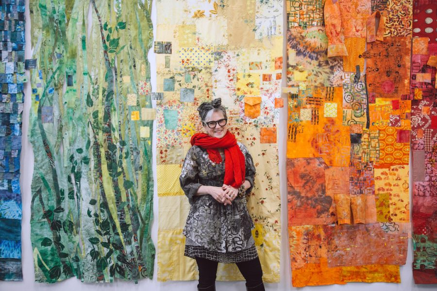 Matching the colorful, patterned background of textiles behind her, artist Merill Comeau stands centered smiling at the camera with a big red scarf.