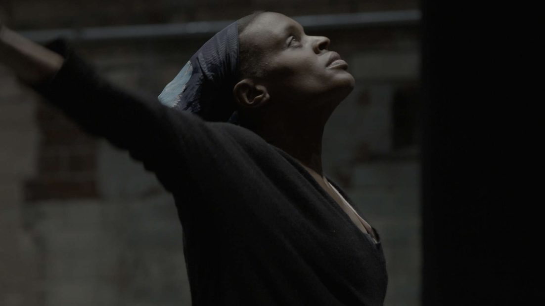 A closed in and profile angle of performer Okwui Okpokwasili, a dark skinned woman wearing black and a headscarf, with her arms stretched wide and looking upward.