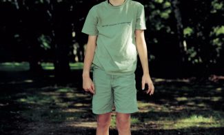 A color photograph of a light-skinned young boy in grayish-green standing in a wooded park.