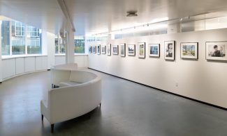 A gallery space with windows at one end, an s-shaped white sofa, and a dozen framed photographs hanging on a long white wall.