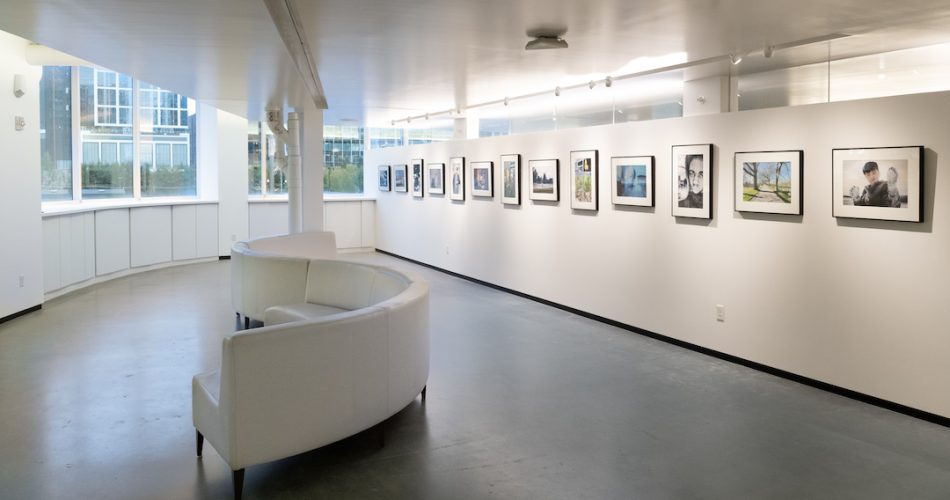 A gallery space with windows at one end, an s-shaped white sofa, and a dozen framed photographs hanging on a long white wall.