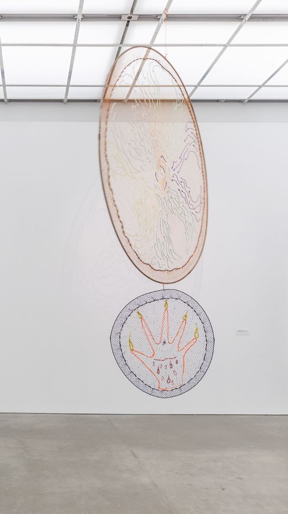 A hanging sculpture made of two fishing nets stretched around two metal rings with colorful, embroidered illustrations
