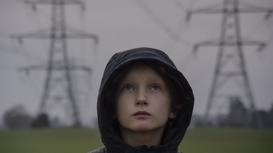 A film still shows a boy of about 9 or 10 in a hoodie facing the camera but looking to the sky. Behind him are two TV towers.