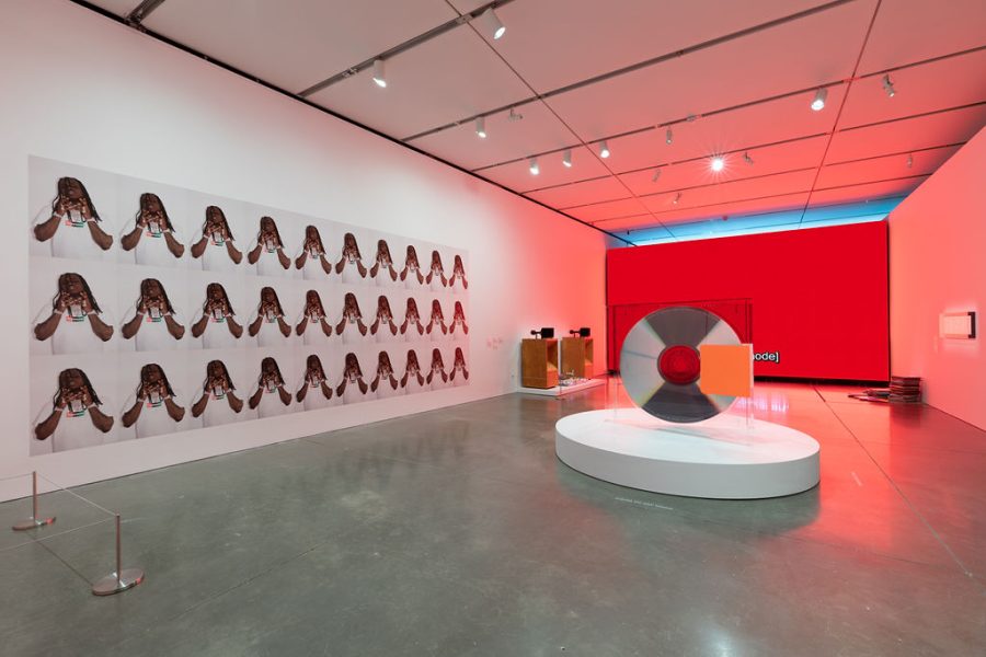 A gallery with a giant standing CD in a case, a lit up red wall, and an image repeated in rows of a dark-skinned man with long dreadlocks. 