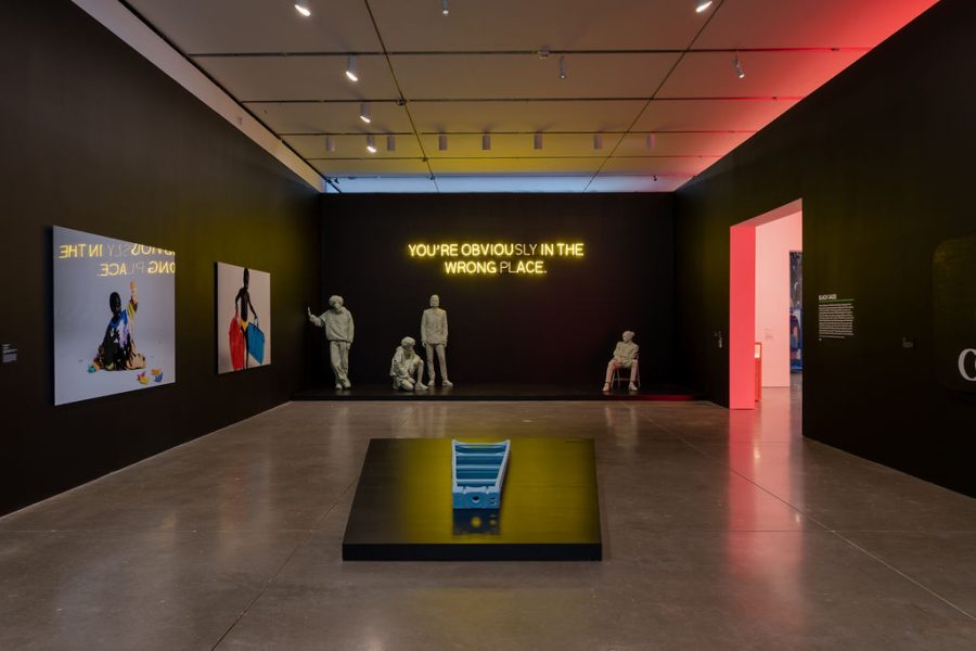 A gallery with a light blue ladder laying on a black plinth, a black wall with the text 