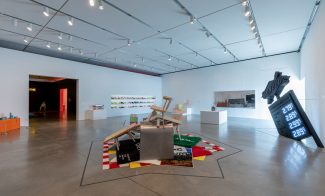 A gallery with a heap of rugs and furniture in the middle, a sculpture resembling a tilted all-black gas station sign on the right, and three shelves of sneakers in the background.