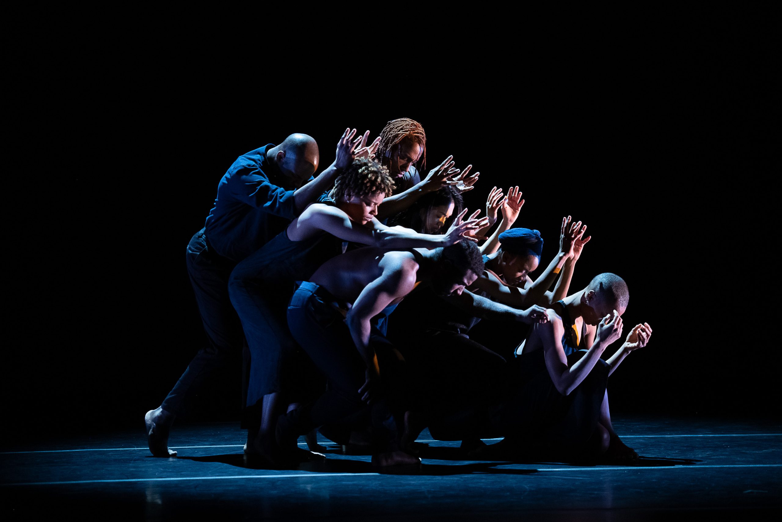 Stage performance of a mass of people grouped together behind one person, lit in dramatic lighting.
