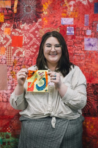 A visitor holding up her quilt square creation in front of a red-patterned tapestry background wall.