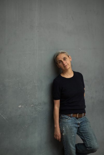 Anne Teresa De Keersmaeker, in a black top and blue jeans, leans against a gray wall.