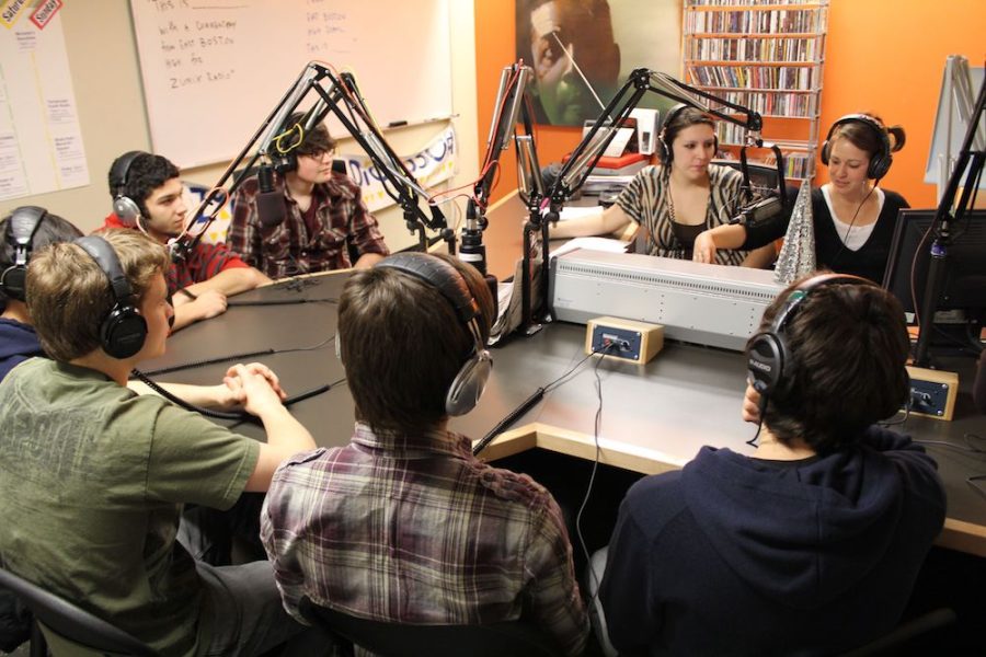 Eight young people wearing headphones gather around a series of microphones and a sound board in a radio studio.
