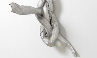 A sculpture of a knotted, tubular form mounted on a wall and mettalic silver in color.