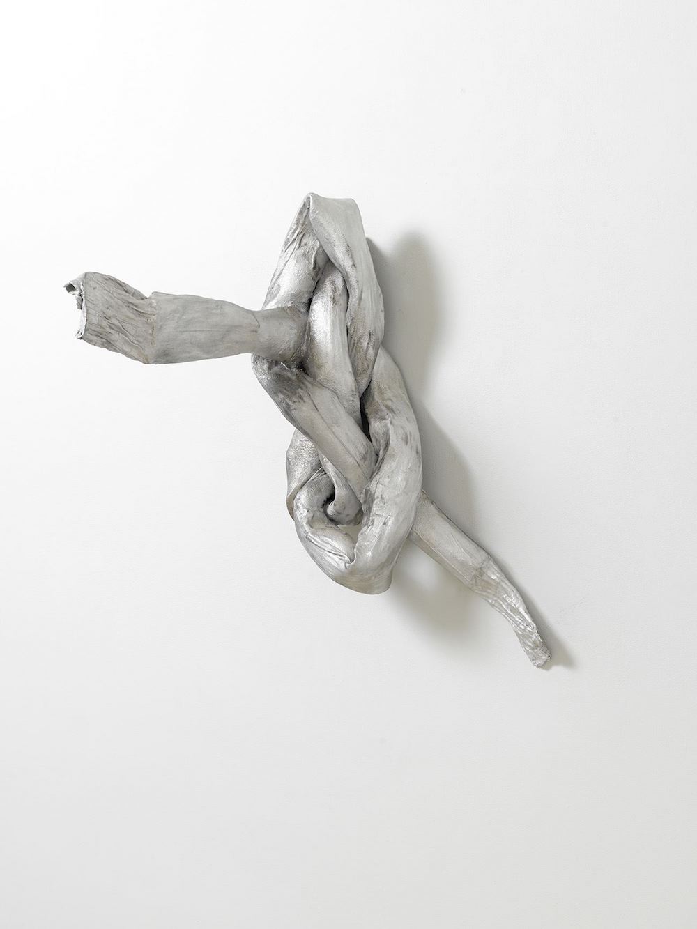 A sculpture of a knotted, tubular form mounted on a wall and mettalic silver in color.