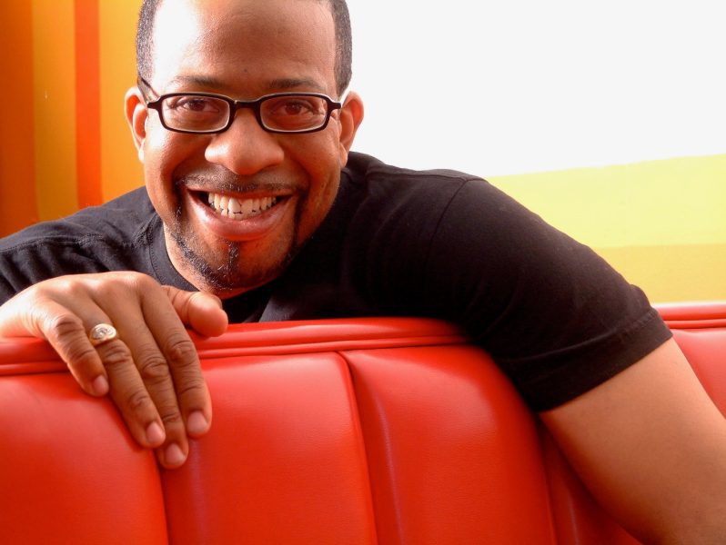 Man with glasses and medium dark skin tone smiles, draped over diner-style seat