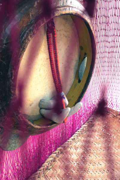 Two sculpted fingers protrude from what appears to be a barrel top or tambourine, positioned vertically. Around that is a magenta-colored net and a rattan surface.