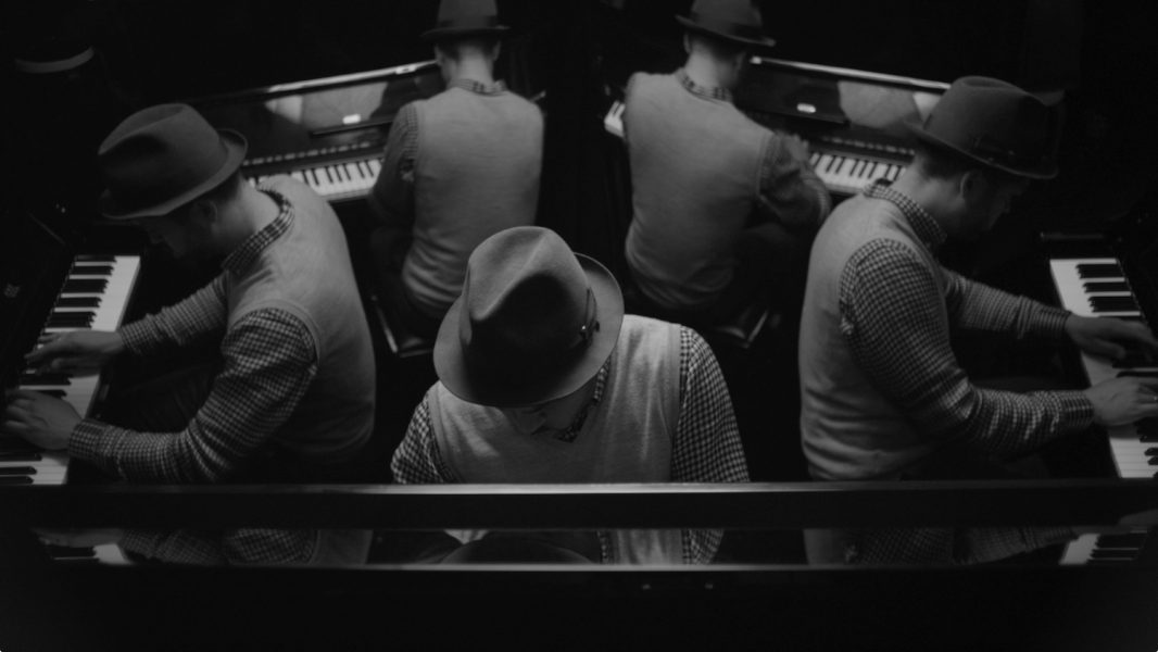 A black and white photographic image shows five men playing piano, facing away from each other, in an image that appears to be reflected.