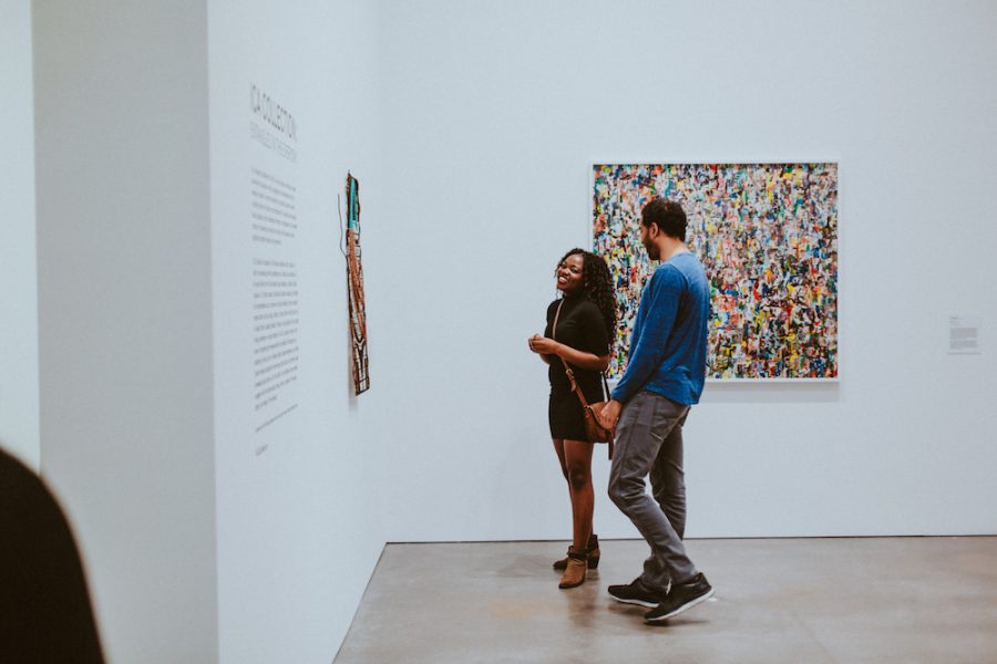A man and woman look toward the left, at an artwork on the wall. She is smiling. A colorful square artwork is behind them.