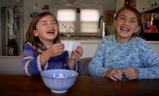 Two children smiling and sitting at the dining table in their home, with one holding up a piece of paper drawn from a ceramic bowl.