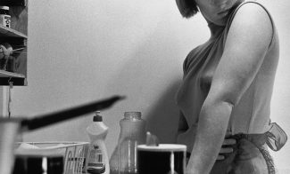A black-and-white photograph of the artist posing in a small, cramped kitchen, looking over her shoulder. She wears a sleeveless top and dramatic eye makeup and the image is cropped to include dishes and dish detergent but cut off the top of her head.