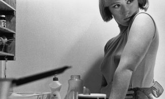 A black-and-white photograph of the artist posing in a small, cramped kitchen, looking over her shoulder. She wears a sleeveless top and dramatic eye makeup and the image is cropped to include dishes and dish detergent but cut off the top of her head.