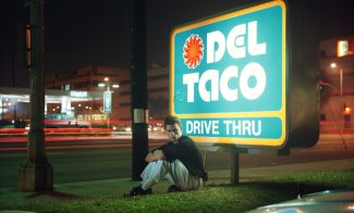 A color photograph of a young, light-skinned man sitting in front of a sign that reads "DEL TACO DRIVE THRU."