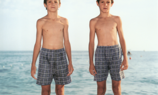 A color photograph of two light-skinned young boys in plaid swimsuits standing side-by-side at the edge of the water on a rocky beach.