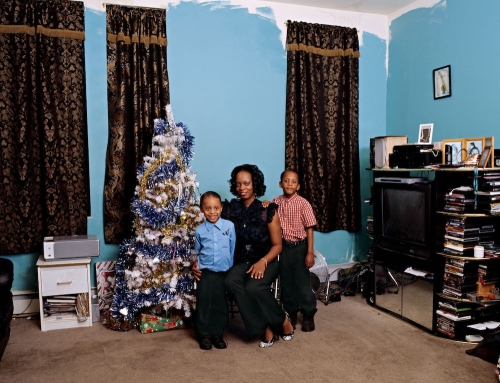 A medium-dark-skinned woman and two children pose in a domestic space with a tinsel Christmas tree and walls painted bright blue.