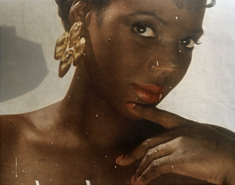 Scratched up reproduction of a vintage-looking photo of a woman with dark skin, gold hanging earrings, and matching coral lips and nails looking at the camera from a slight angle