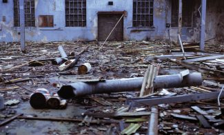 A color photograph of an industrial yard covered in debris with a dilapidated building in the background.