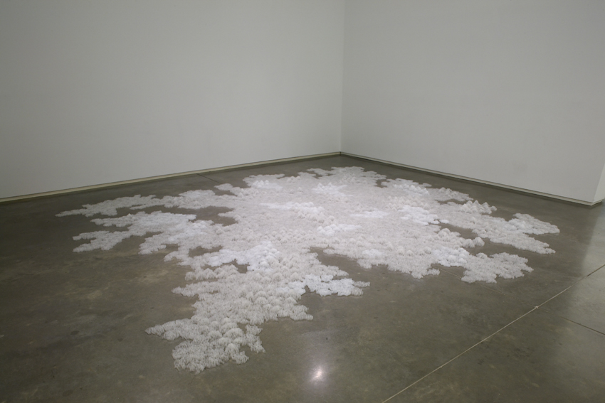 A sculpture of a low white form composed of looped Scotch tape extending across a cement floor.