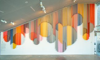 A monumental hanging wall sculpture made from bands of colorful coated mesh fabric which drape in various lengths to create series of interlocking circular forms, and installed in an empty spacious lobby.